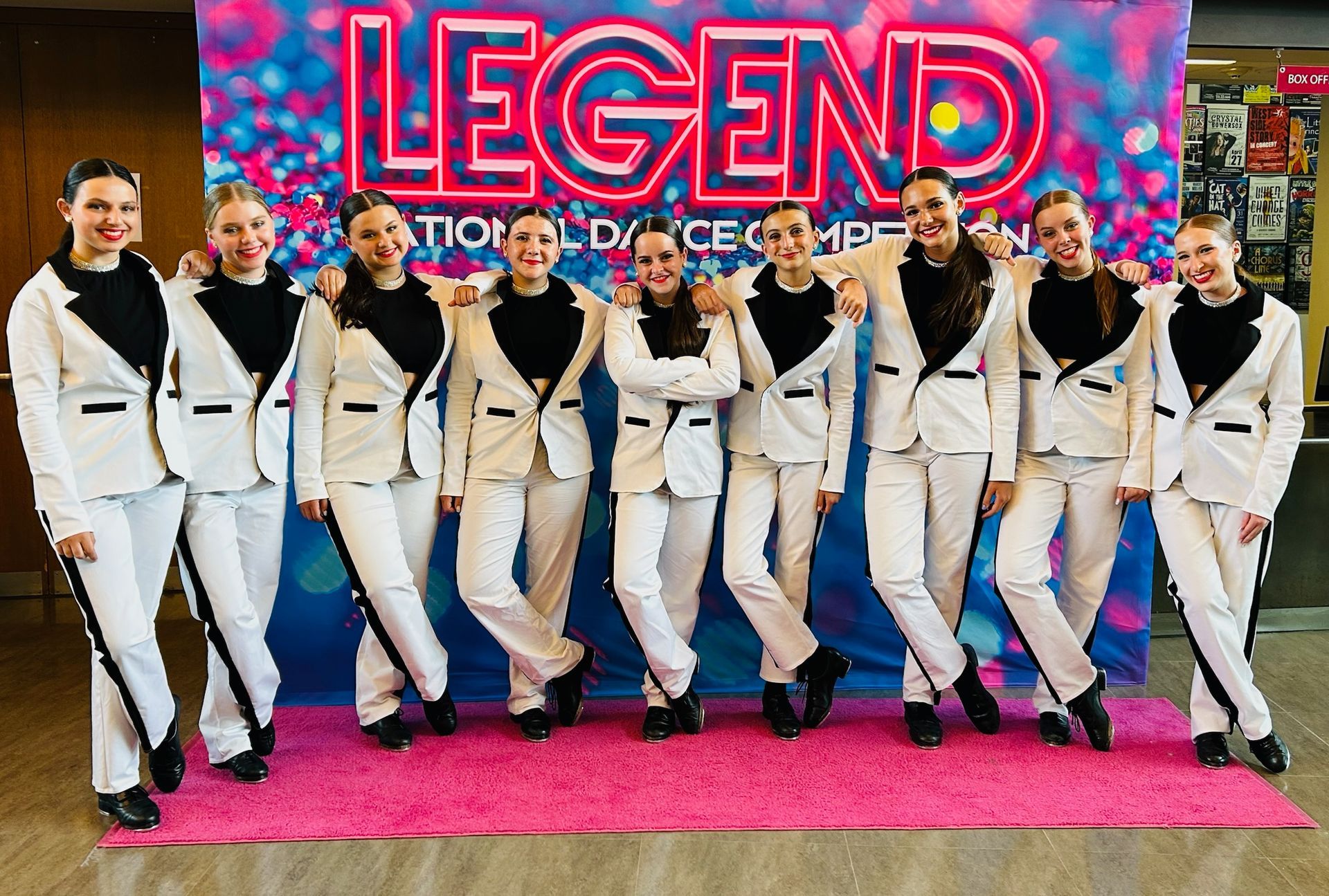 A dance team poses at the Legend National Dance Competition. The group of eight young women wear white suits, black tops, and stand in front of a colorful backdrop.