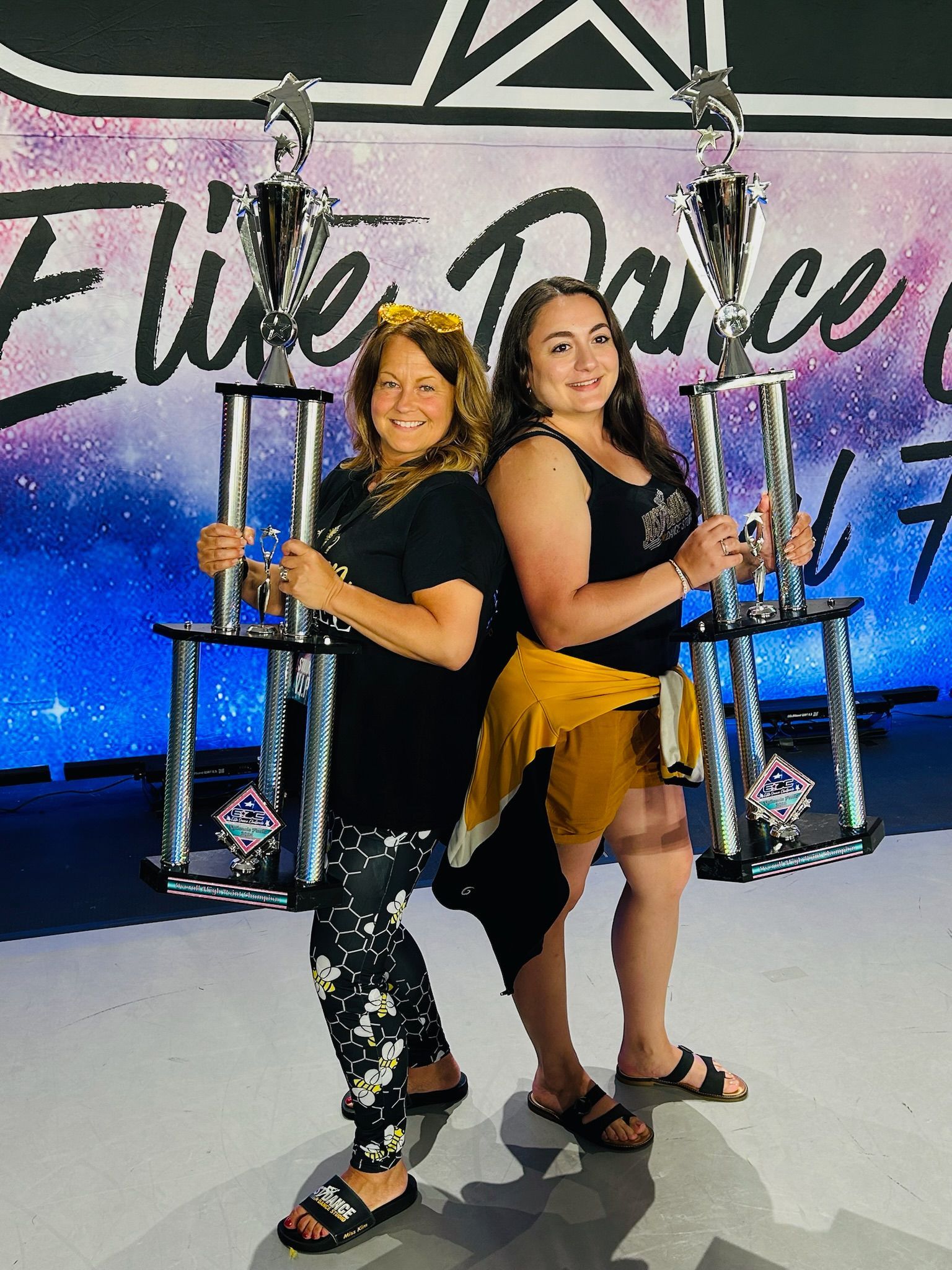 Two women stand back-to-back, holding large trophies. They are in a dance studio with a starry background.
