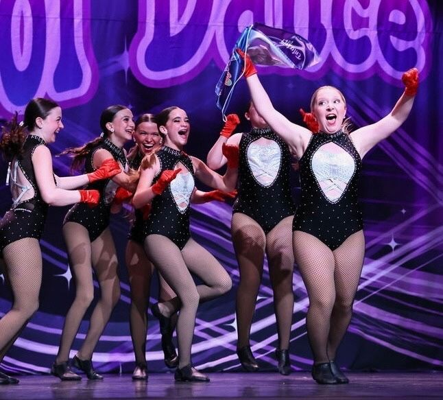 A dance team in black leotards with red gloves celebrates onstage, one holding a flag. Purple backdrop.