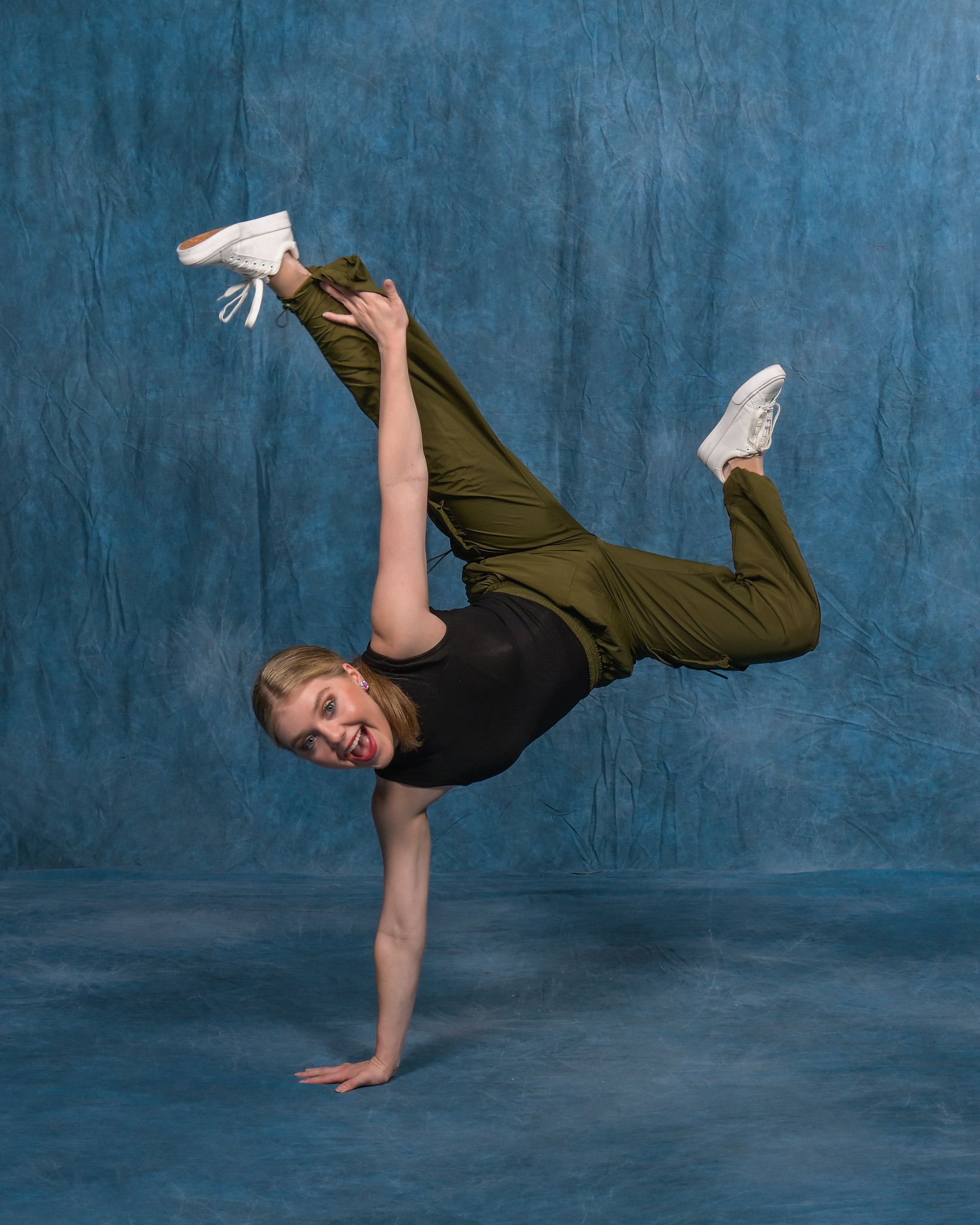 A young woman in athletic attire balances on one hand, legs extended mid-air in a dance pose, smiling, with a blue background.