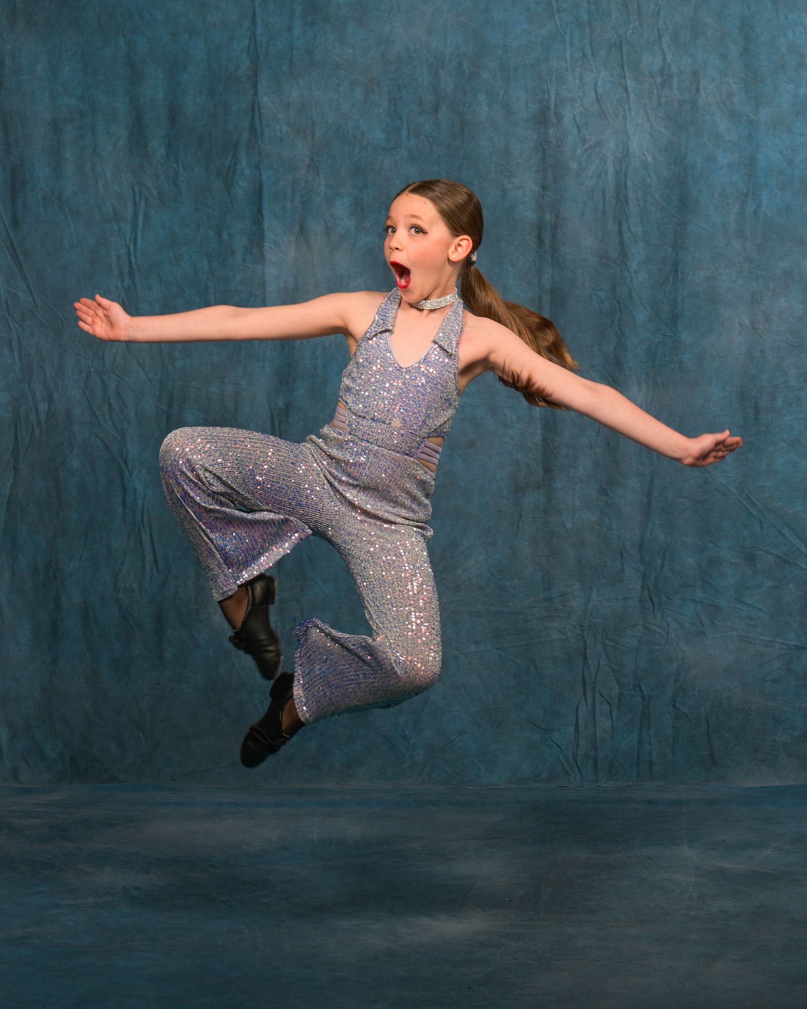 Girl in a sparkly jumpsuit leaps in the air against a blue background, mouth open in a surprised expression, arms outstretched.