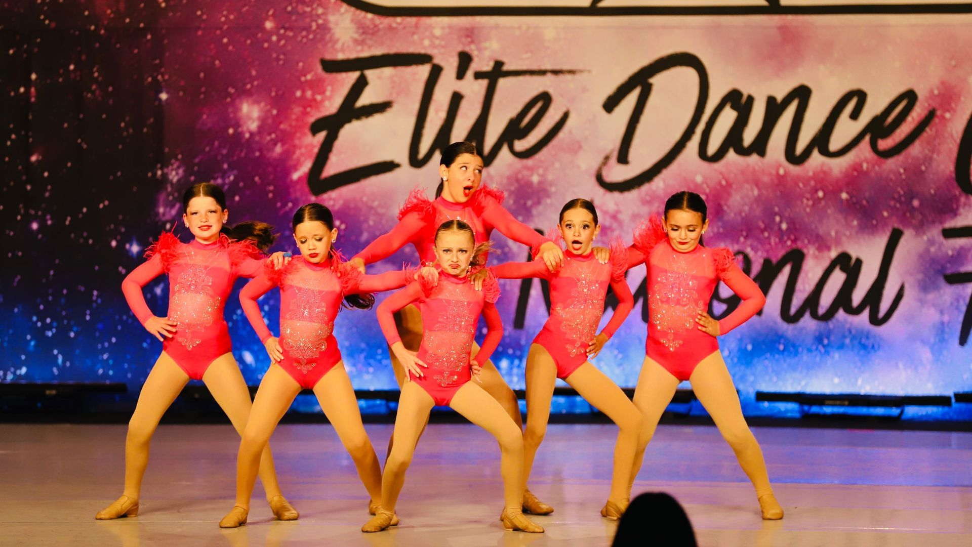 Six young dancers in red costumes strike a pose on stage with a cosmic backdrop.