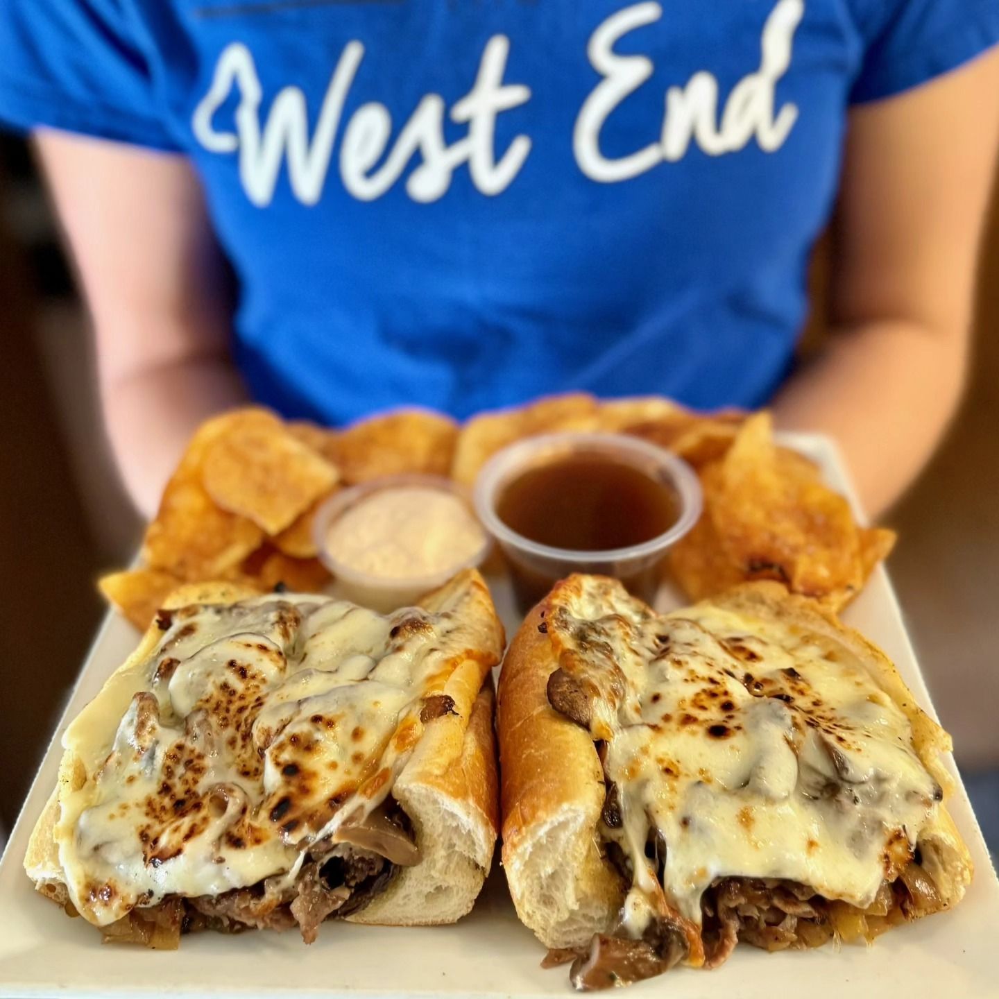 Person holding a plate with two French dip sandwiches, chips, and dipping sauces.