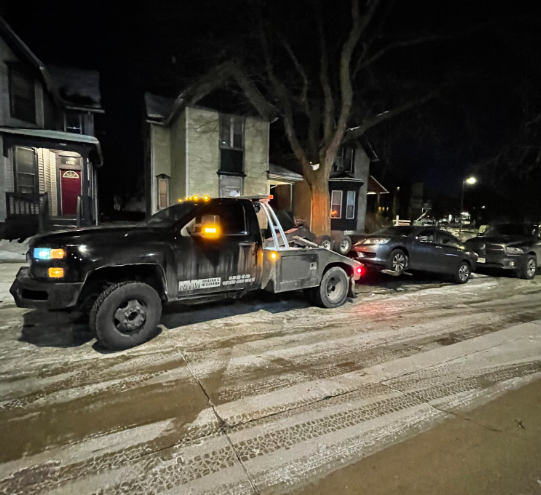 Tow truck towing two cars on a snow-covered street at night.