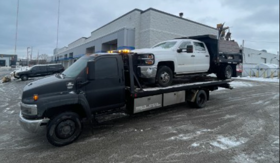 Tow truck hauling a white pickup truck on a cloudy day. Building in background, snow on the ground.