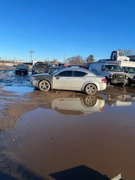 Silver car in muddy puddle; junkyard setting with other vehicles and clear sky.