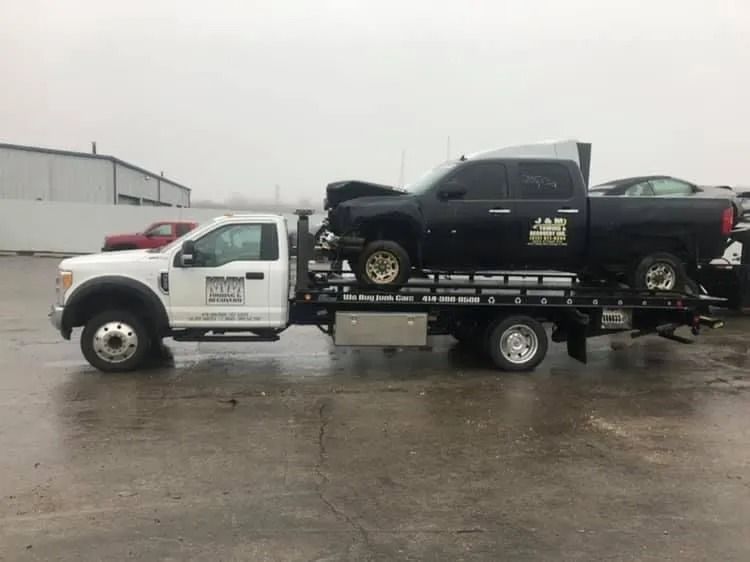 White tow truck carrying a black pickup truck with damage on a rainy day.