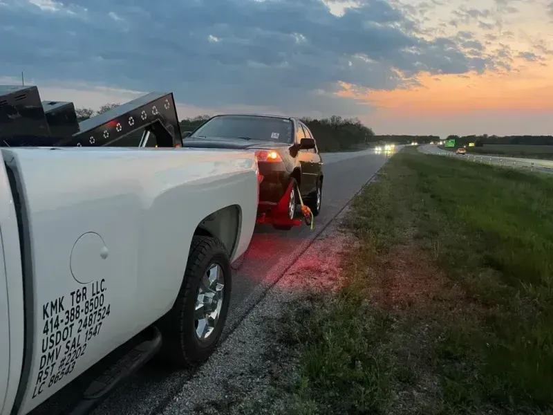 Tow truck towing a dark SUV on the shoulder of a highway at dusk, with red taillights illuminated.