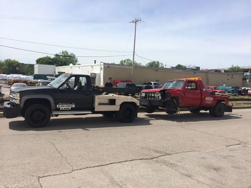 Tow truck towing a damaged red truck on a sunny day in a parking lot.