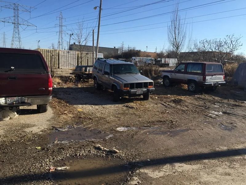 Dirt lot with three SUVs and a pickup truck parked near a fence and power lines on a sunny day.