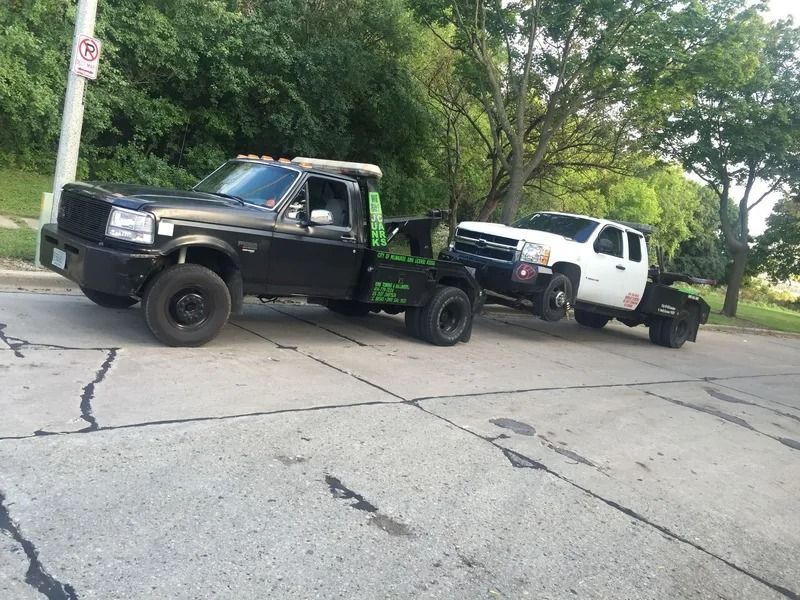 Black tow truck towing a white pickup truck on a cracked street, both facing right.