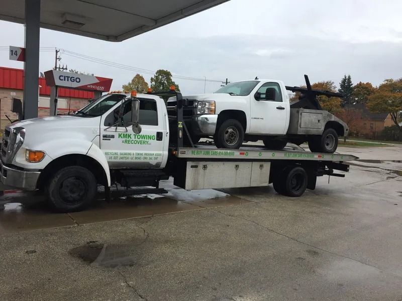 White tow truck towing a white pickup truck at a gas station under a canopy.