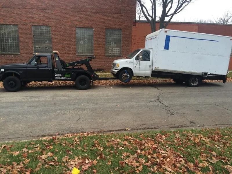 Black tow truck towing a white box truck on a street, autumn leaves on the ground, brick building in the background.