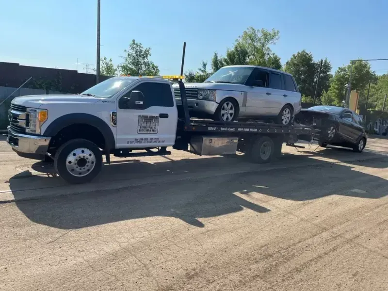 Tow truck carrying two vehicles on a sunny day. A silver SUV is on the flatbed, and a dark car is being towed.