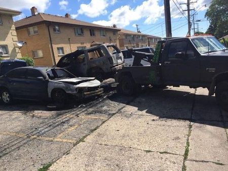 Damaged vehicles on a residential street; smoke marks on pavement, burnt SUV, dark truck.