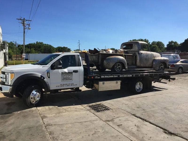 Tow truck carrying a rusted vintage pickup truck on a sunny day.