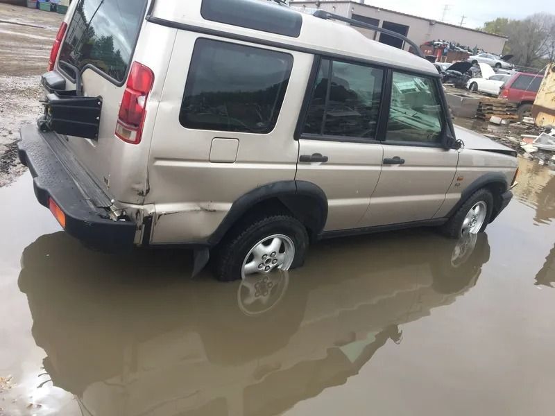 Tan Land Rover Discovery partially submerged in muddy water at a salvage yard, with damage to rear.