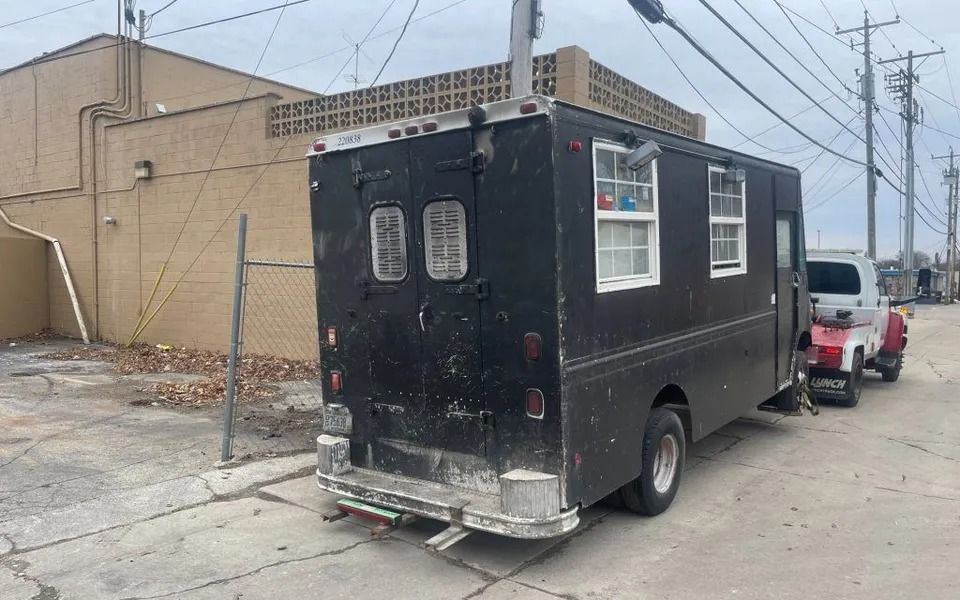 Black food truck parked on a paved street next to a brick building and a white truck.