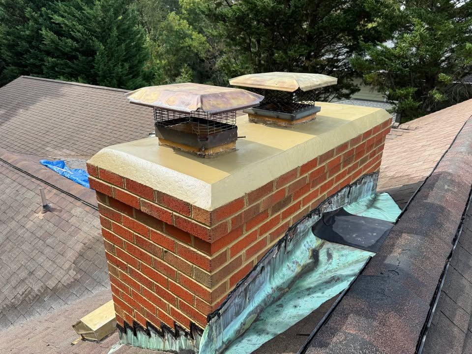 Brick chimney with two flues, metal caps, and weathered roof in a sunny setting.