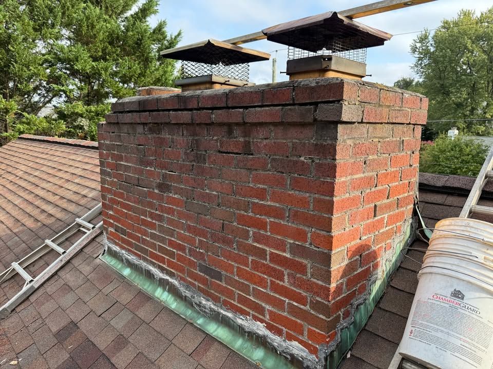 Brick chimney on a shingled roof, with chimney caps, a ladder, and a paint bucket visible.