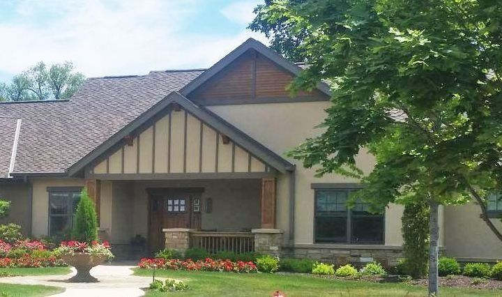 Beige house with a covered porch and brown roof, green lawn, and flowering garden.