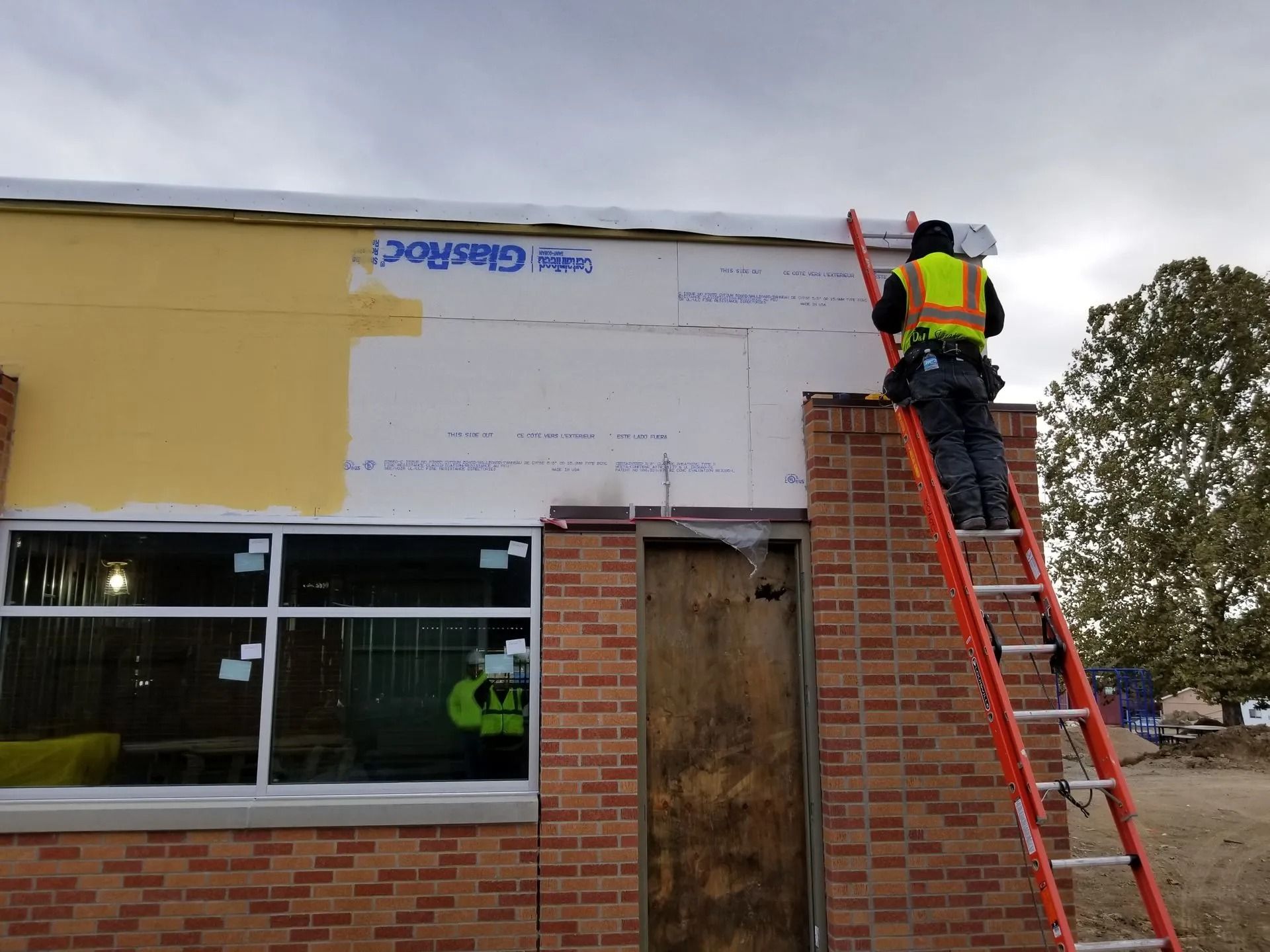 Construction worker on a ladder, installing white wall panels on a brick building under a cloudy sky.