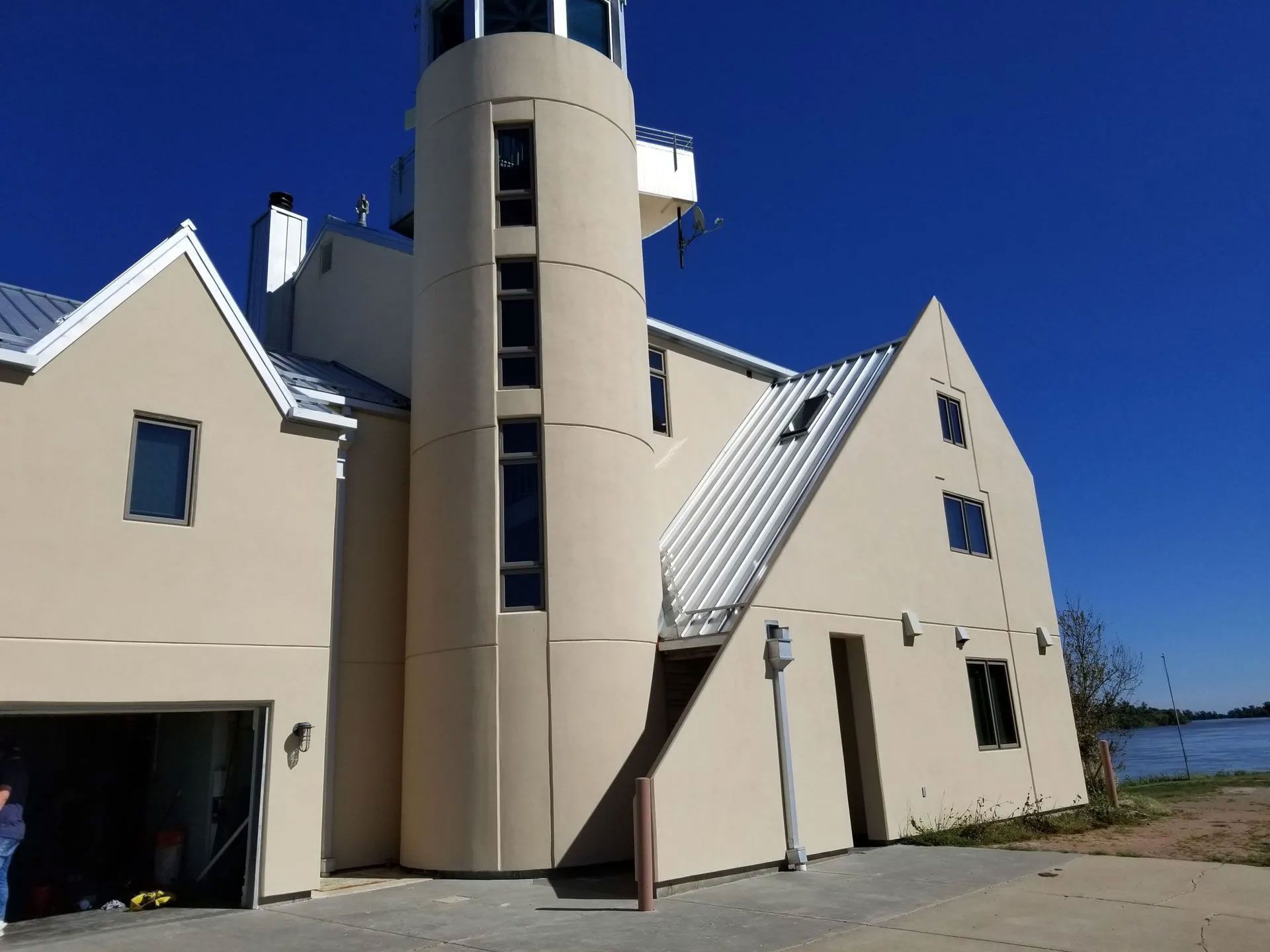 Beige building with a cylindrical tower and a peaked roof, set against a bright blue sky near a body of water.