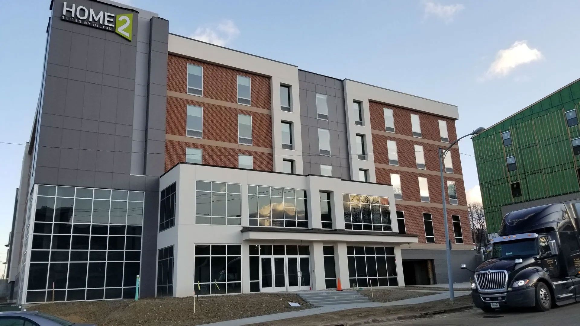 Exterior of a Home2 Suites hotel building with gray and brick facade. A semi truck is parked in the foreground.