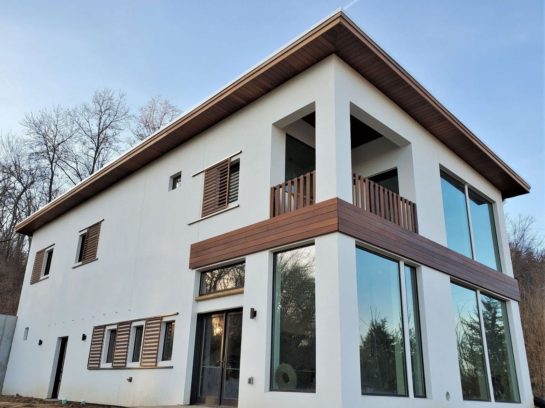 Two-story white house with brown accents, large windows, and balcony, set against a blue sky and trees.