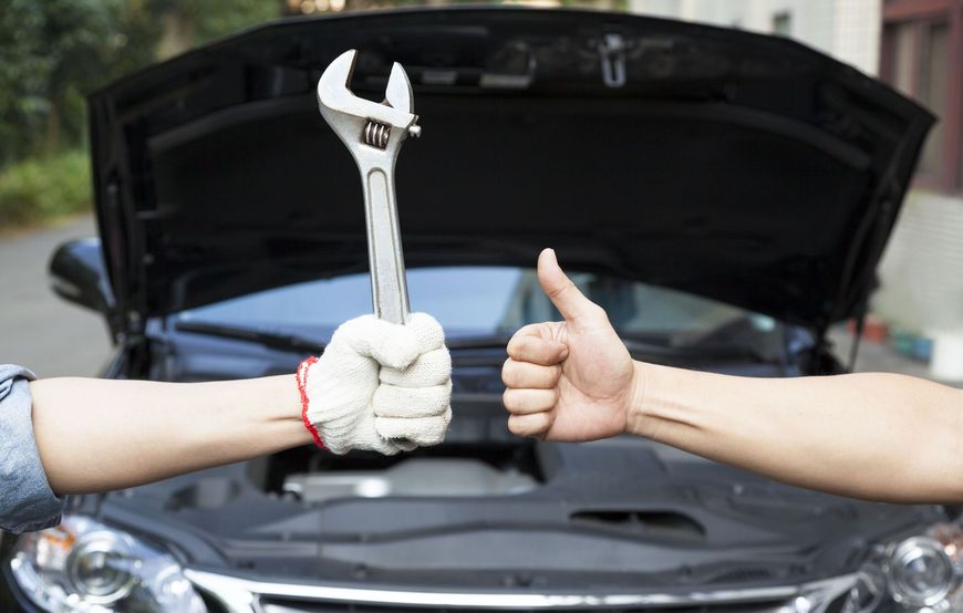 Hands holding wrench in front of car with open hood; one giving a thumbs-up.