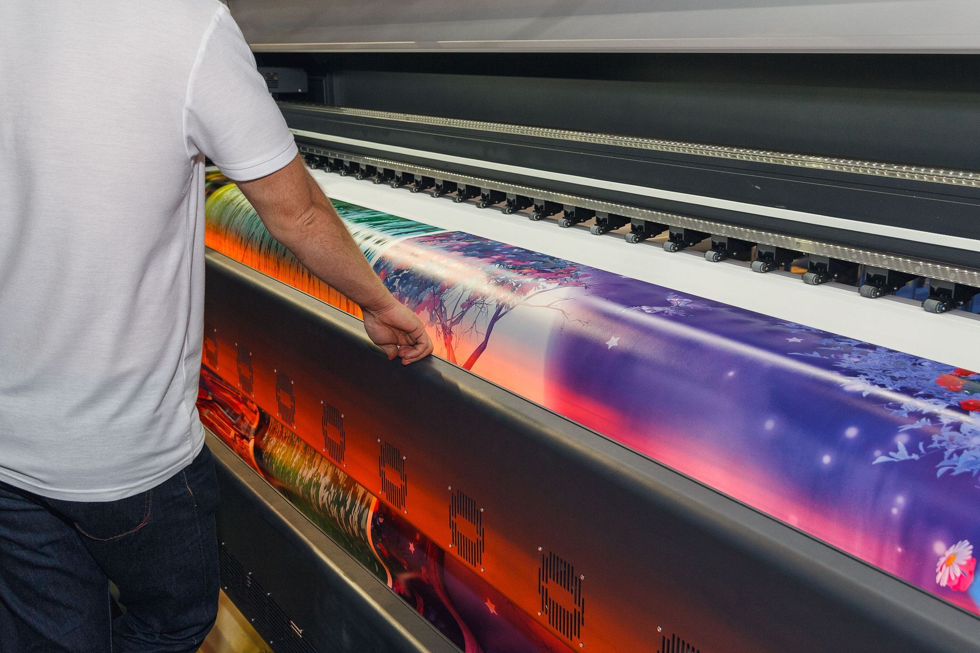 A person operates a large-format digital printer, guiding a vibrant, colorful printed graphic through the machine.