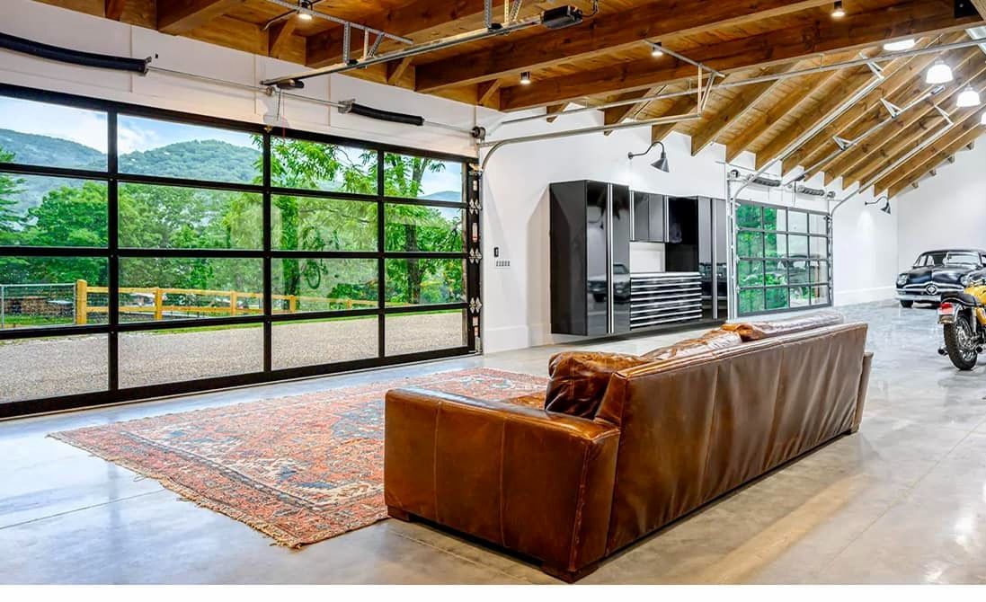 Spacious modern interior with glass garage door overlooking a green landscape. A leather sofa sits on a rug, with kitchen and vehicles visible.