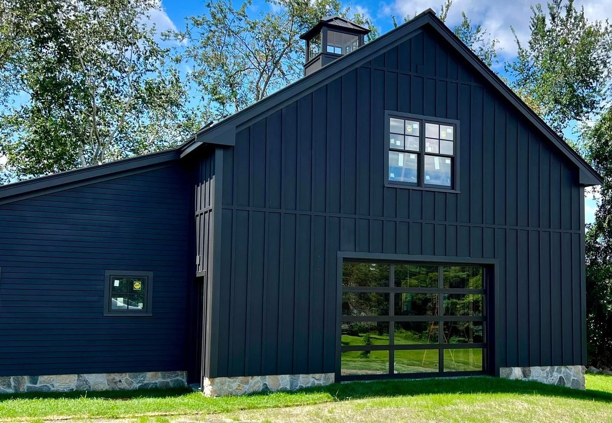 Dark blue barn with a glass garage door, small square windows, and a cupola on the roof. Situated on a green lawn.