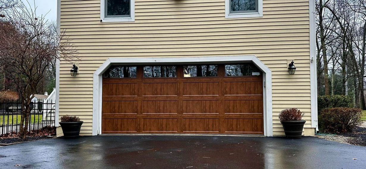 A brown garage door framed by light yellow siding. Two small windows are above. A paved driveway and two potted plants are in front.