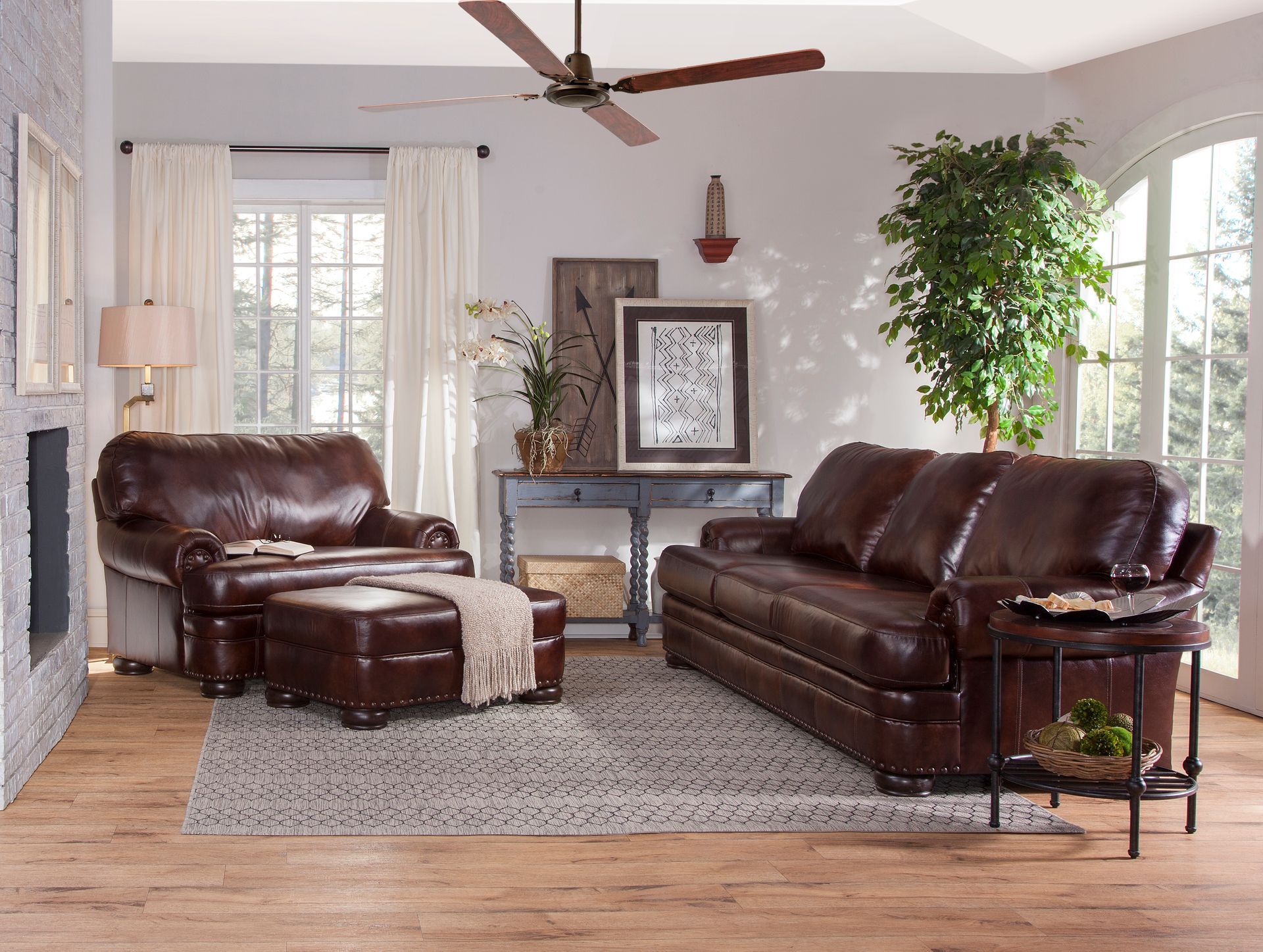 A living room with a brown leather couch , chair , ottoman and ceiling fan.