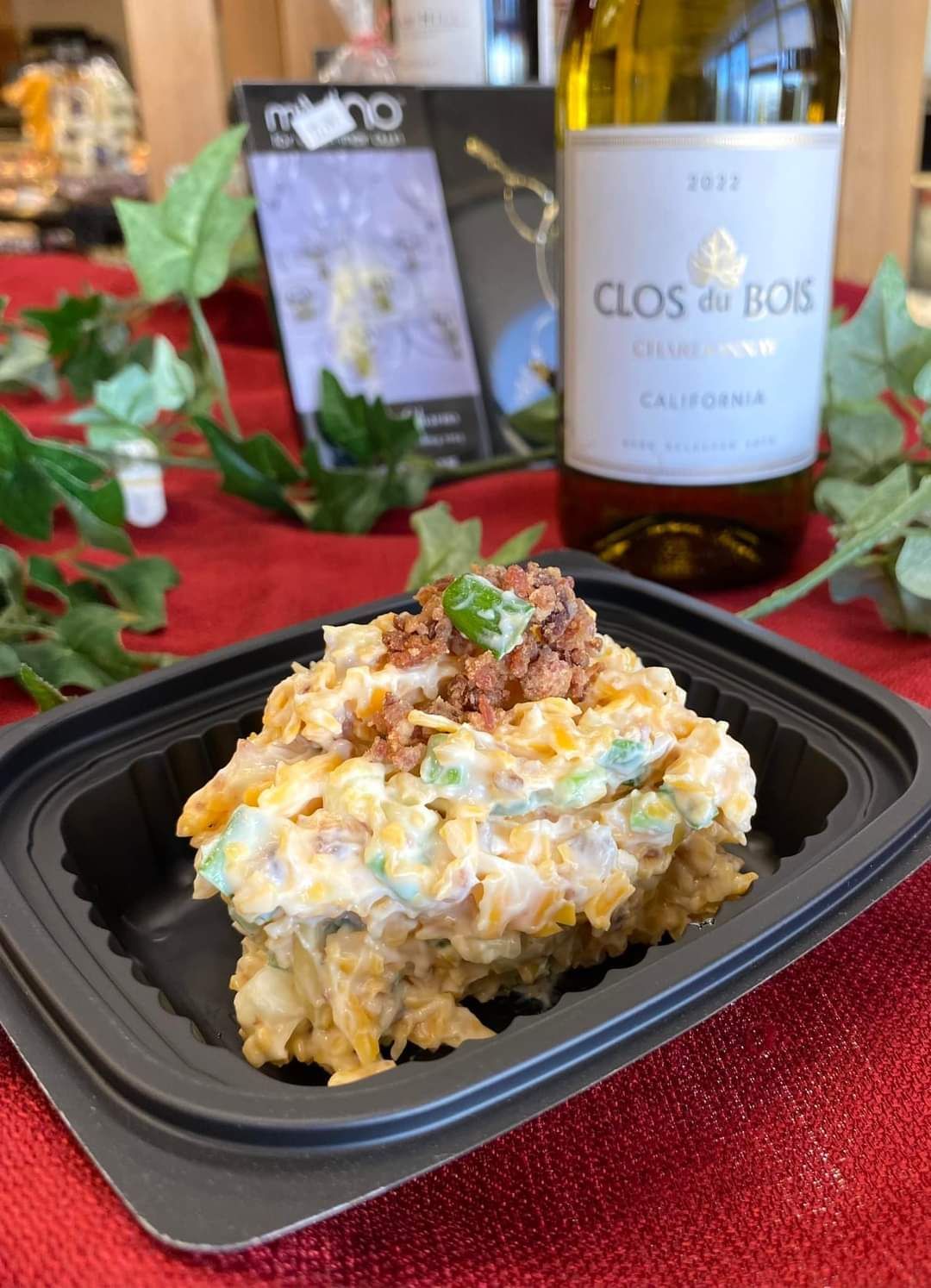 Creamy potato salad in a black takeout container beside a wine bottle and holiday greenery