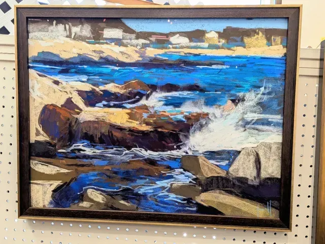 Framed seascape painting with blue waves, rocky shore, and cloudy sky on a store shelf.
