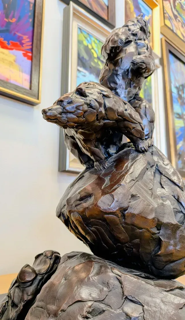 Bronze sculpture of a seated figure in a gallery, with colorful framed paintings behind it.