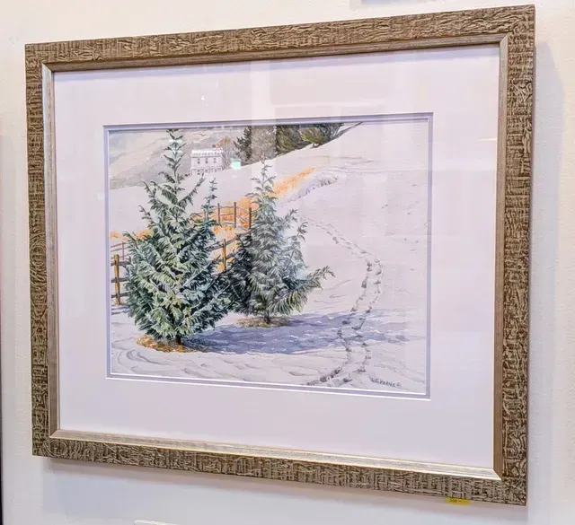Framed watercolor of snowy evergreen trees in a gold decorative frame hanging on a wall.