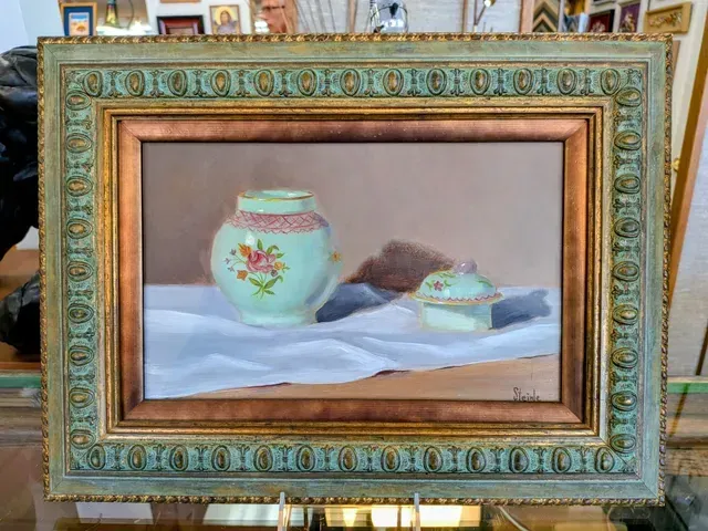 Framed still life painting of a white vase and teacup on draped cloth against a pink background.