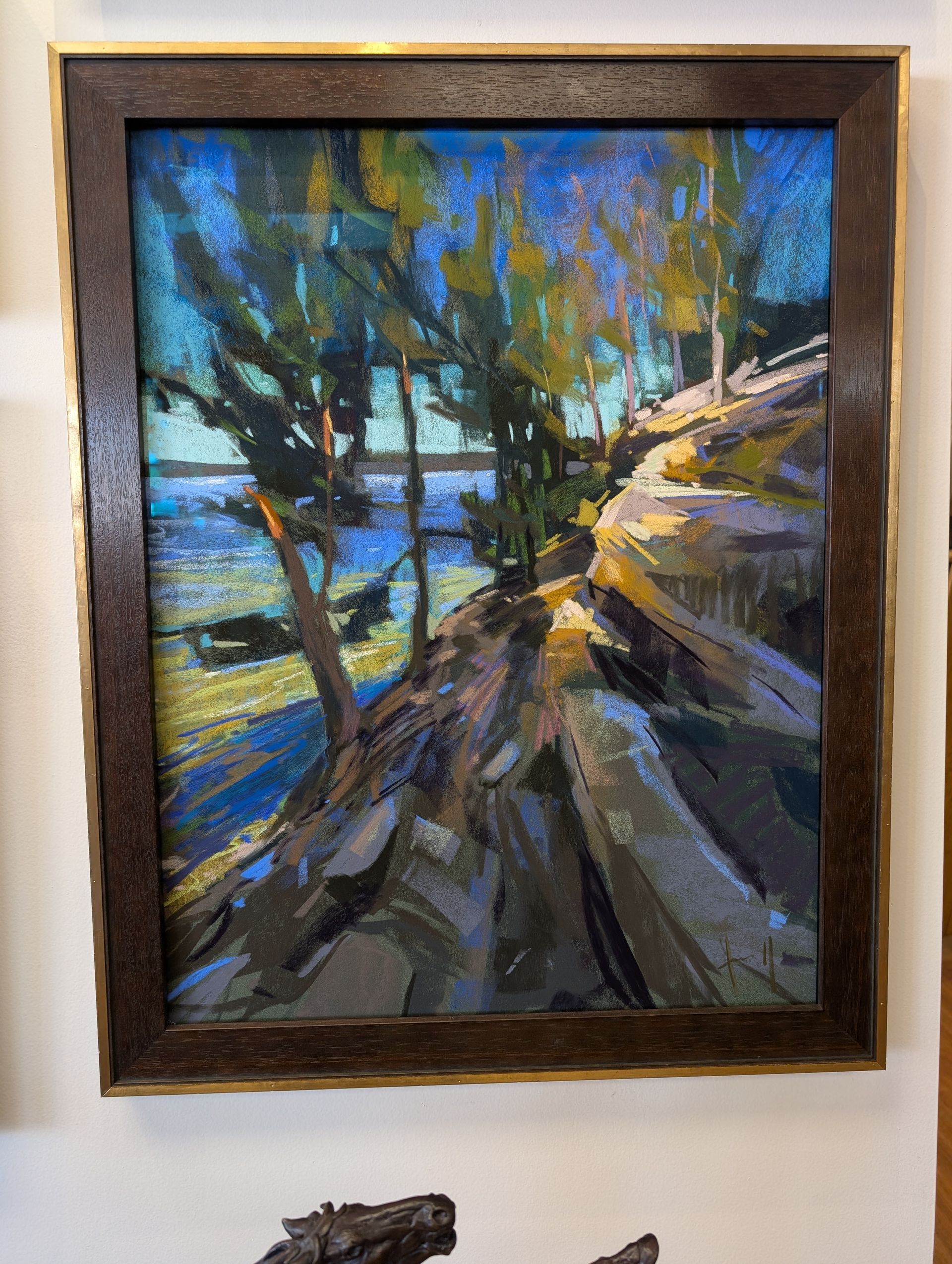 Framed impressionist painting of a sunlit path by blue water and trees, hanging on a wall.