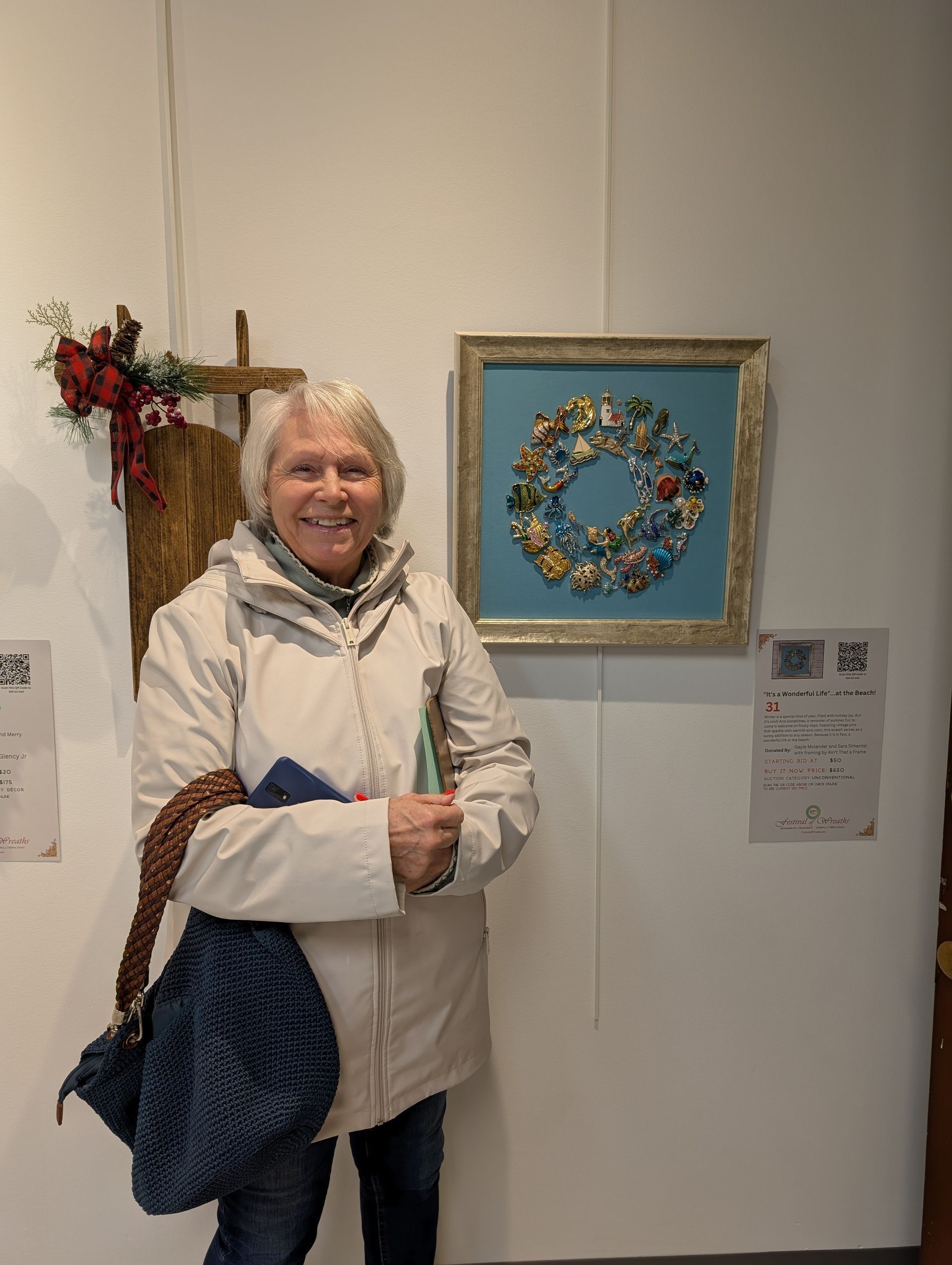 Woman in a white coat smiling beside a framed floral wreath on a gallery wall.