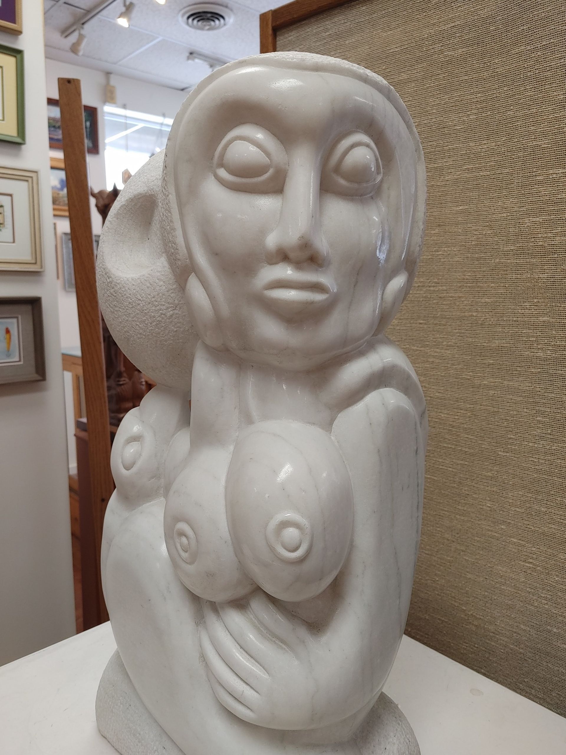 White abstract sculpture of a seated female figure in a gallery, with closed eyes and stylized breasts.