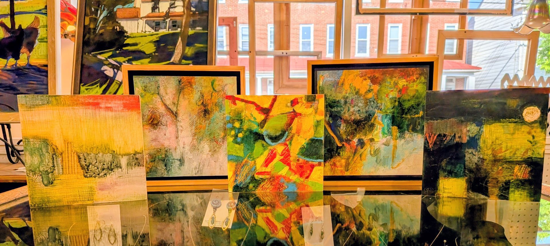 Abstract colorful paintings displayed in a row before a glass wall with reflections and brick buildings behind.