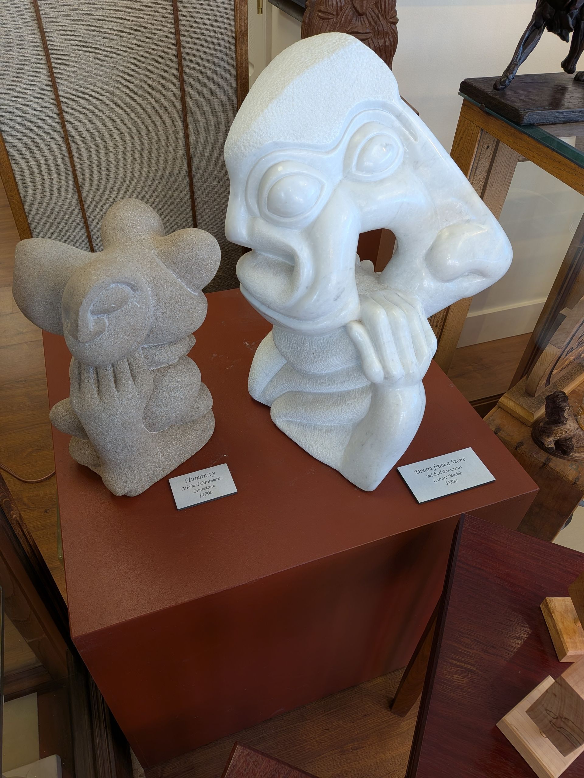 Two sculpted figurines on a wooden pedestal, one gray and one white, displayed indoors with small labels.
