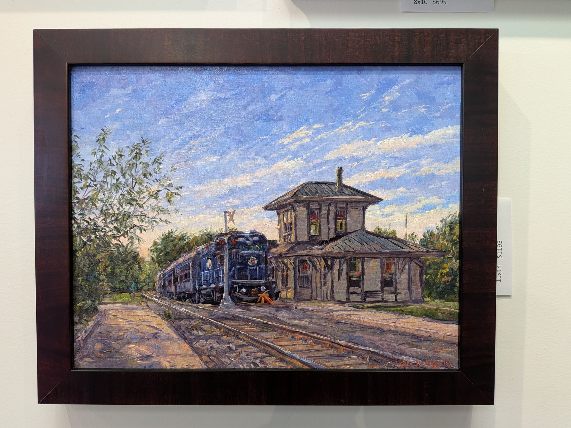 Framed painting of a steam train beside a rustic station under a blue sky with clouds.