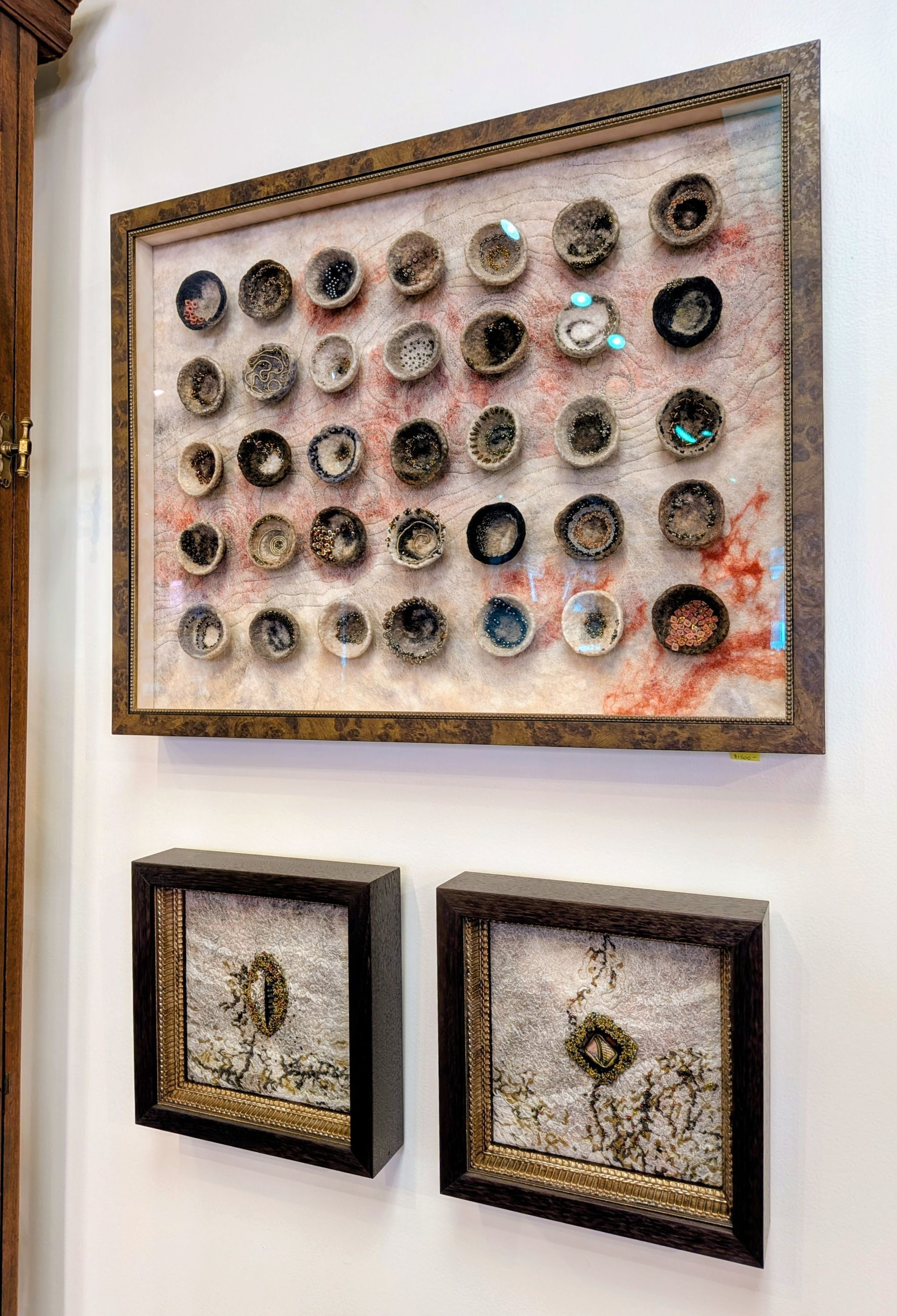 Framed wall art with many circular objects and two smaller framed pieces below on a white wall.