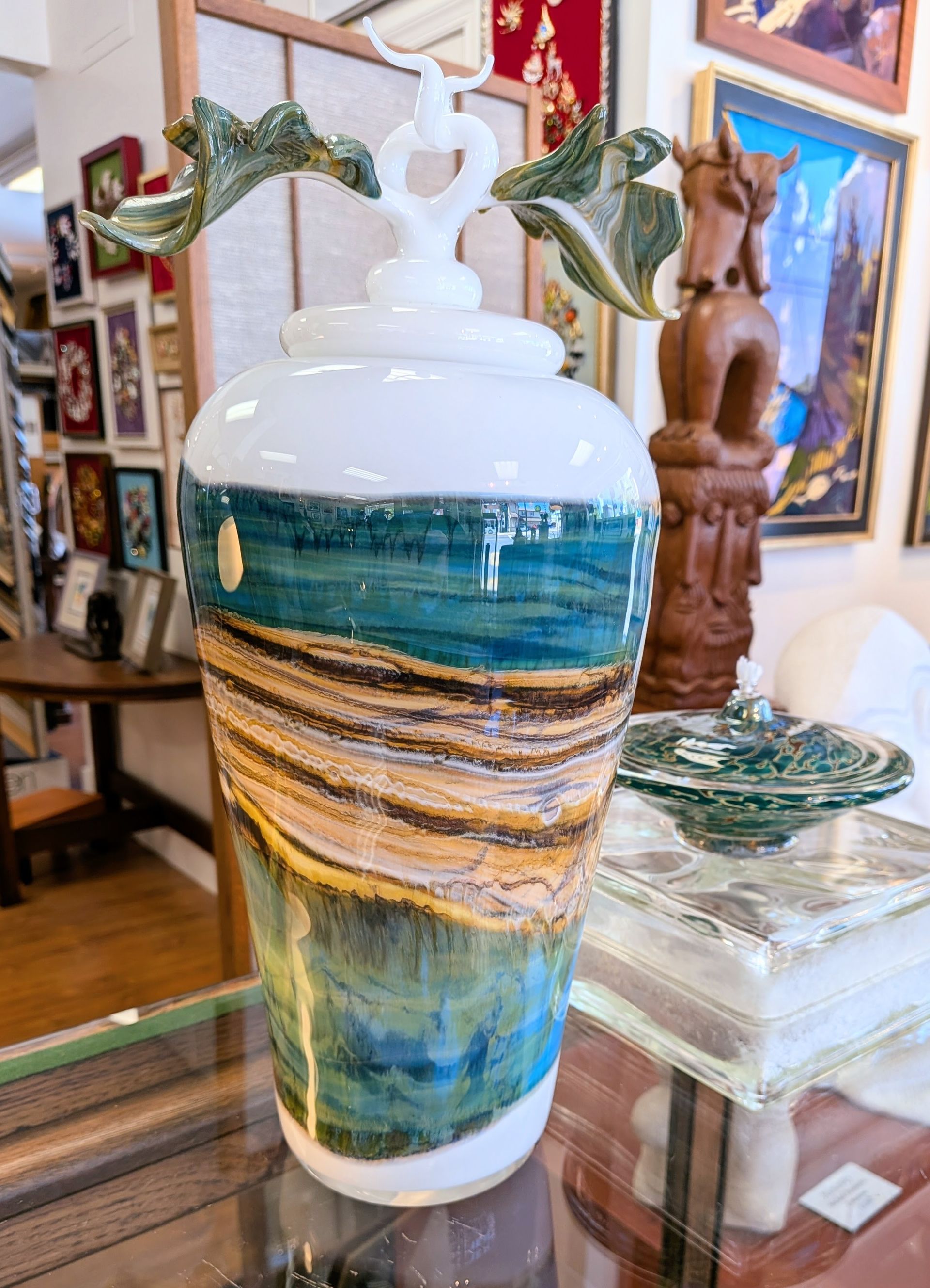 White ceramic vase with blue, gold, and green swirls, topped with decorative leaves, on a glass table in an art-filled room.