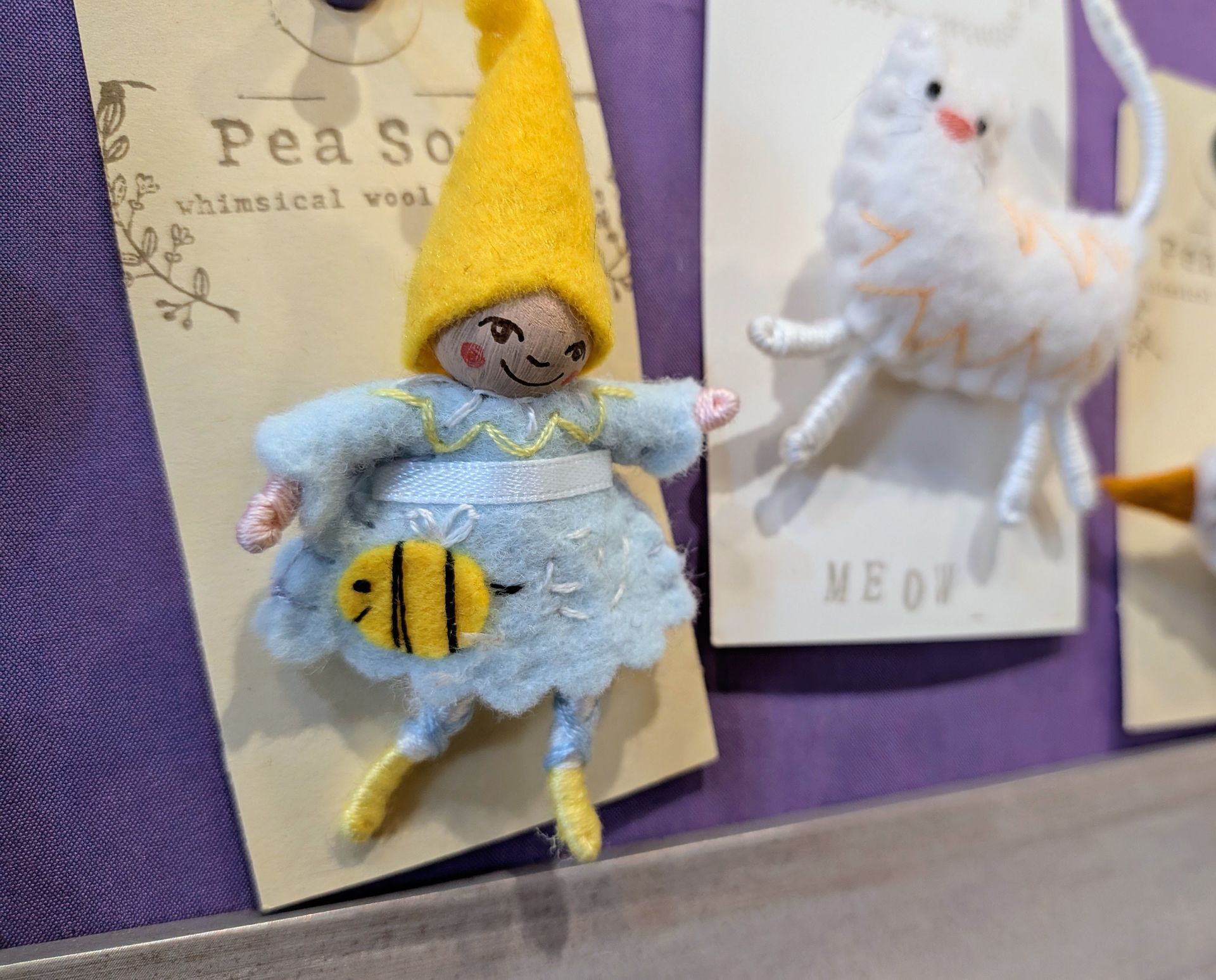 Small felt bee costume figure with a yellow hat, displayed on a purple board beside a white plush toy.
