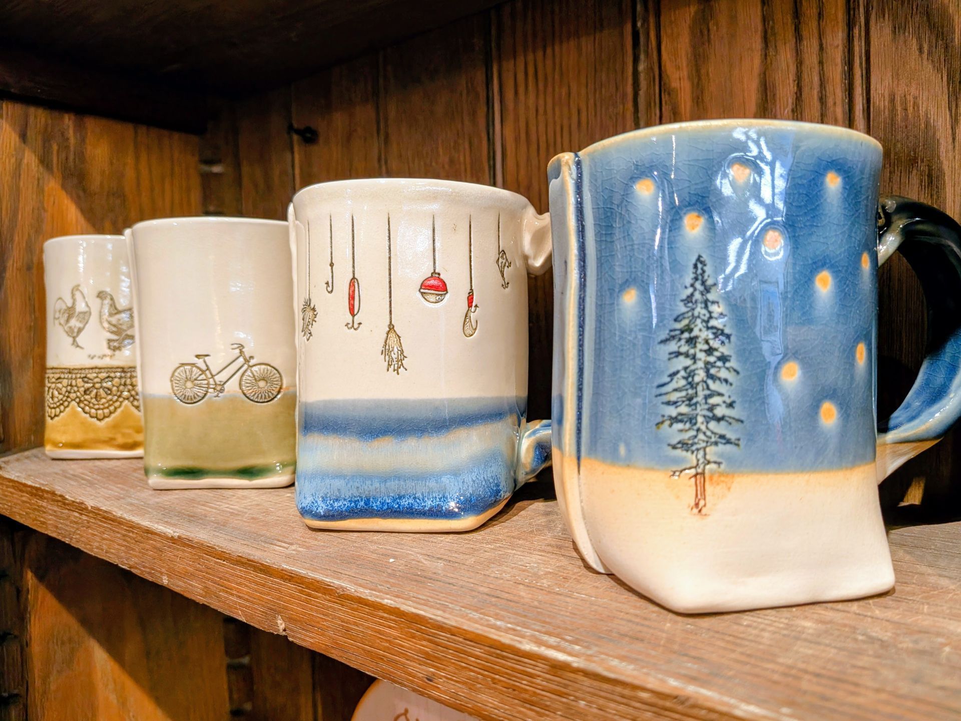 Hand-painted ceramic mugs with winter scenes and blue accents displayed on a wooden shelf.